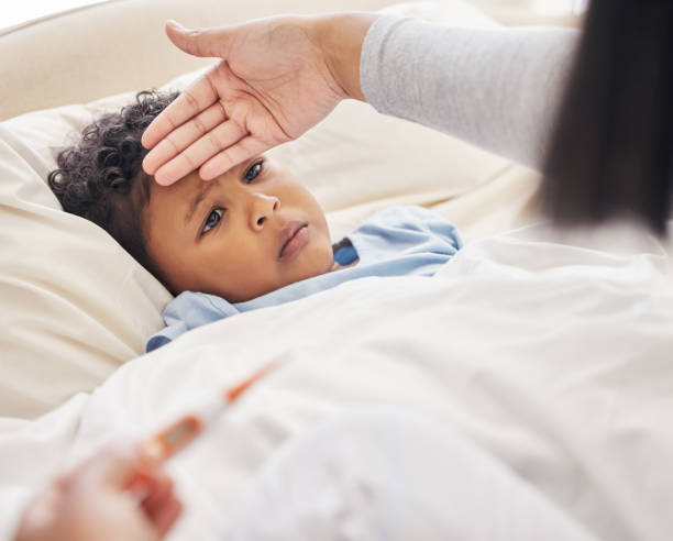istock Sick little boy lying in bed while mother takes his temperature with thermometer and feeling forehead. Mother taking care of child with fever 1398824672