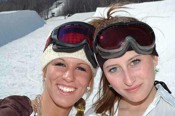 Two young athletic snowboarder women posing together for headshot smiling into the camera in front of the halfpipe at Okemo Mountain resort in Vermont, USA.