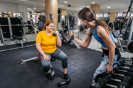 Young female gym instructor coaching a young plus size woman in the gym. Lifting weights while instructor is taking a picture using a smart phone.