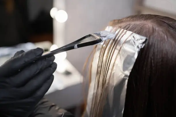 Close-up of a strand of female hair on a special sheet of foil, the master applies hair dye with a brush. The process of highlighting by a professional master in a beauty salon. Life style