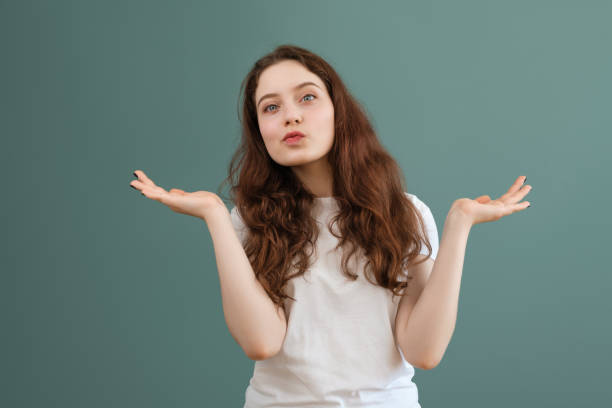 Teenage brunette girl in white t-shirt spreads her hands, on teal background. I don't know, confused gesture. stock photo