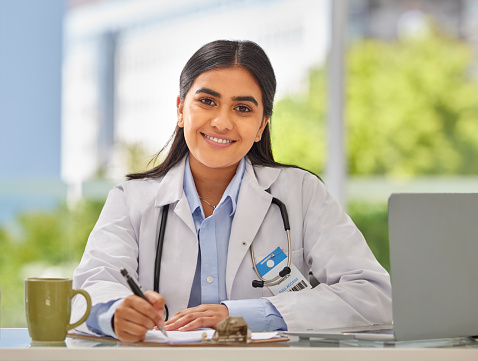 Portrait of a young indian doctor wearing a stethoscope sitting in a office writing a prescription while sitting at her desk. Smiling female gp or physician writing in medical file in clinic