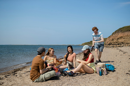 Group of mixed ethnic teens sitting on a beach together at Holy Island in the North East of England in summer. They are eating a packed lunch and one man is running behind his girlfriend, getting ready to scare her.
