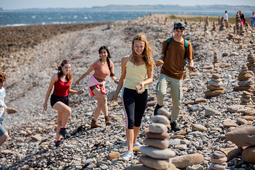 Wide shot of a group of mixed ethnic teens building pebble stacks together while at a beach together at Holy Island in the North East of England in summer.