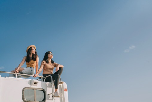 Asian Chinese Lesbian couple enjoying scenic view on top of campervan outdoor road trip during weekend morning