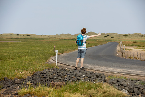 Young man standing at the side of a non-urban road holding out his thumb, hitch hiking at Holy Island in the North East of England in summer.
