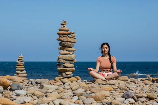 Wide shot of a young woman sitting meditating with her eyes closed beside a pebble stack she has built while at a beach at Holy Island in the North East of England in summer.