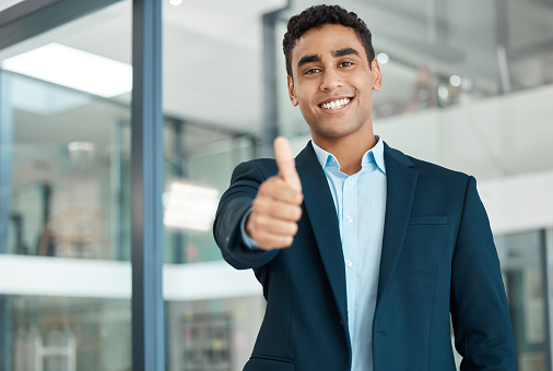 Young happy mixed race businessman showing a thumbs up standing alone at work. One pleased hispanic businessperson smiling while showing support with a thumbs up in an office