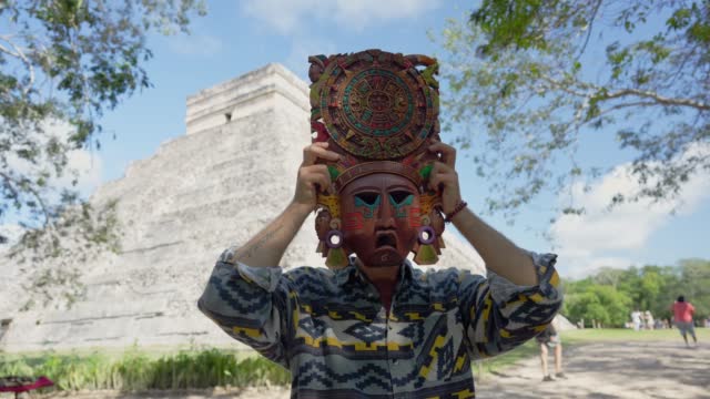 Man holding Mayan mask at his face on the background of Chichen Itza