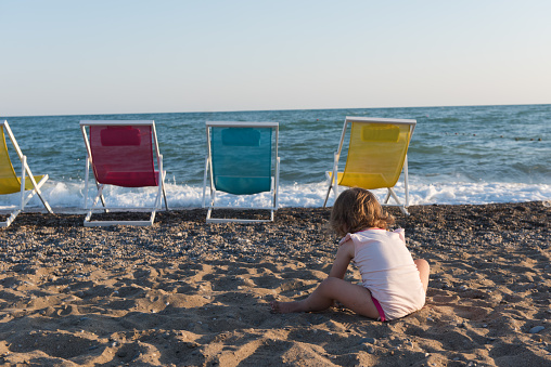 Little girl in sitting on sand and playing with bucket and spade at sea beach in warm sunny summer day. Closeup. Side view. High quality photo