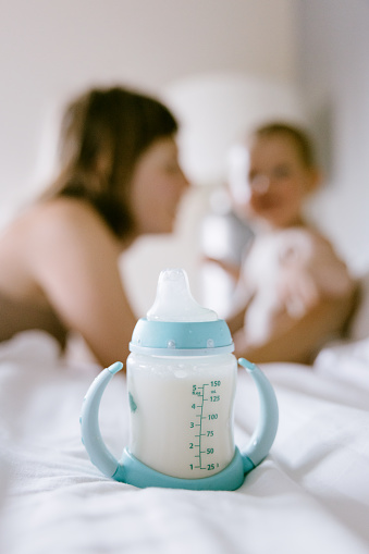 A Caucasian mom plays with her baby son, a bottle of baby milk formula in focus in the foreground.  Shortages of baby formula have caused food security issues for many families in the United States.