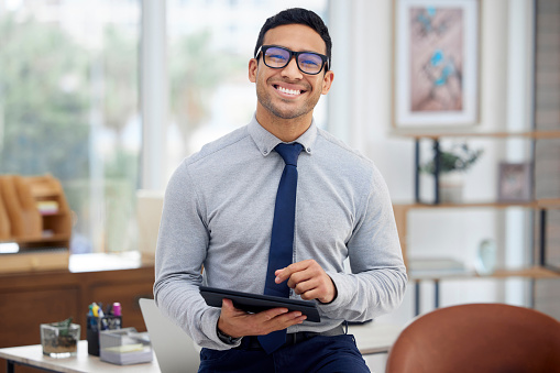 Portrait of a young mixed race businessman holding and using a digital tablet standing in an office at work. One content hispanic male businessperson typing on a digital tablet at work