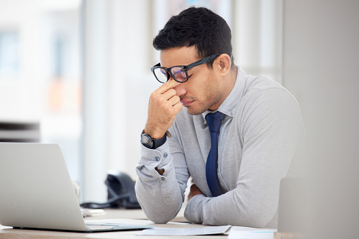 Tired mixed race businessman suffering from a headache while working on a laptop at work. Stressed hispanic male businessperson looking upset in an office