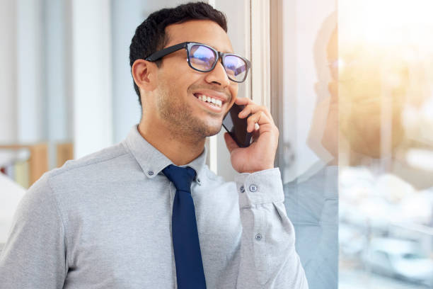 istock Young content mixed race businessman on a call using a phone and  standing in an office at work. One hispanic male businessperson smiling while talking on the phone standing and thinking at a window in an office 1398844478