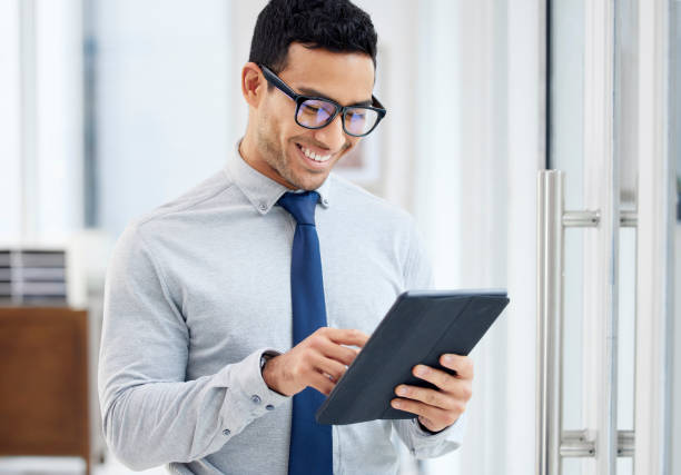 istock Happy mixed race businessman holding and using a digital tablet standing in an office at work. One content hispanic male businessperson smiling while typing an email on a digital tablet at work 1398844483