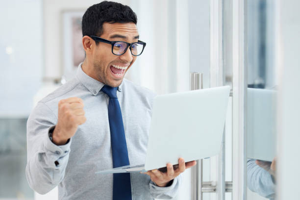 istock Amazed young mixed race businessman standing and working on a laptop in an office at work. Joyful hispanic male businessperson looking shocked while cheering and holding a laptop at work 1398844497