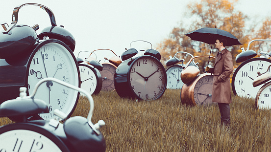 Concept of dealing with issues related to time. Man stands with umbrella outside looking at large collection of big alarm clock on a field.