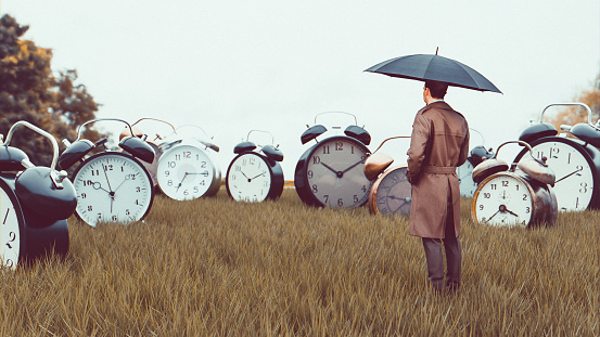 Concept of dealing with issues related to time. Man stands with umbrella outside looking at large collection of big alarm clock on a field.