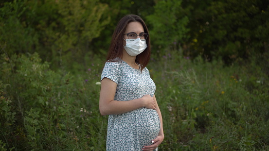 A young pregnant woman in a medical mask stands in nature. The girl in glasses and a dress looks into the camera. 4k