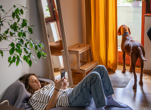 Young woman lying on bag chair at home using smartphone with hungarian pointer dog