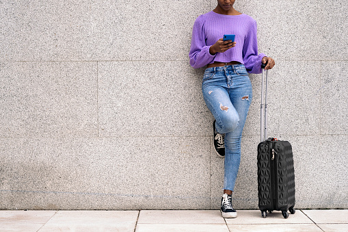 Stock photo of unrecognized afro woman with suitcase using her phone.