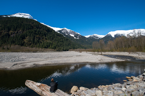Man sitting on a tree log and enjoying the breathtaking view of snow-capped mountains reflecting in the Squamish river in British Columbia, Canada, on a bright sunny day in Springtime.