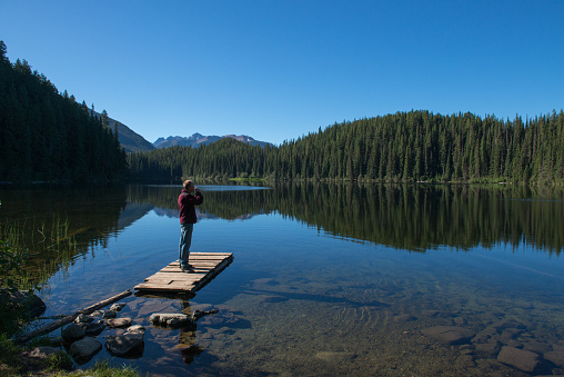 Man starting the day and drinking coffee in early morning, and standing on a wooden dock while watching trees and mountains reflecting in a quiet lake. British Columbia, Canada.