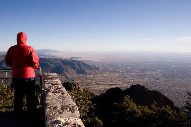 A man in a red jacket looks off the top of a mountain at mountains and a city below.