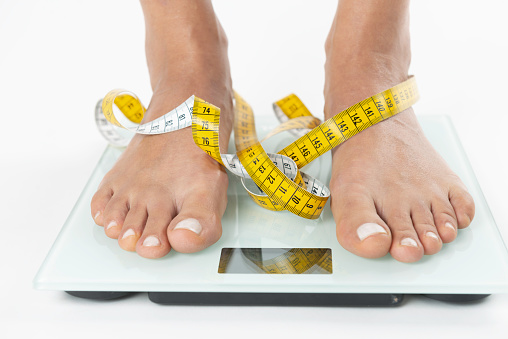 Barefoot female on weighing scale with tape measure on white background.