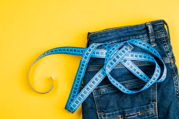 the concept of a healthy lifestyle and diet. blue jeans with a blue measuring tape instead of a belt - loose weight imagens e fotografias de stock