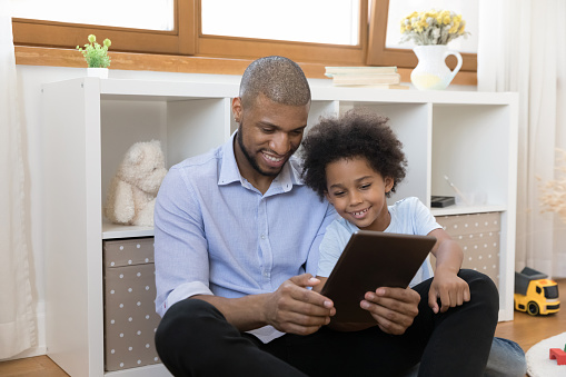 Young African father his cute little 6s son sit in playroom having fun use tablet, enjoy educational amusing app, play video games, watching vlog content, buying on e-commerce retail services concept