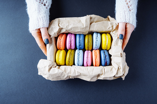 Box of macaroons in female hands on a dark background. View above.