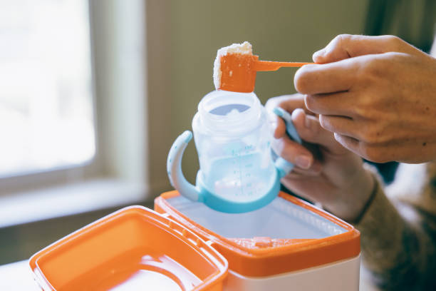 A Caucasian parent scoops infant formula for feeding their baby into a bottle.  Current formula shortage in the United States has many families feeling food insecurity.