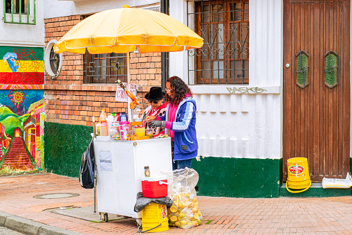 Bogota, Colombia - January 27,2017: A couple of Colombian women sell Juices and snacks at the street corner in the historic La Candelaria in the Andean capital city of Bogota in Colombia in South America. It was in this area that the Spanish Conquistador, Gonzalo Jiménez de Quesada founded the City in 1538. Many of the walls in this area are painted in the vibrant colours of Latin America, or with Street art that may include local tribal legends from the pre-Colombia era.  Photo shot in the morning sunlight on an overcast day; horizontal format.