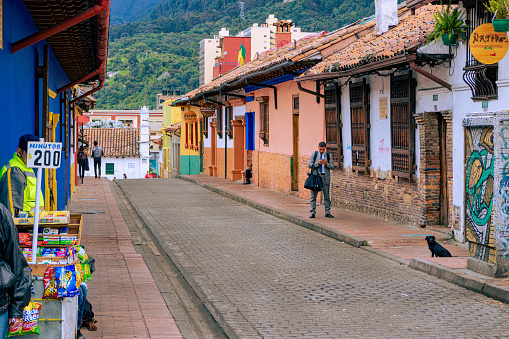 Bogotá, Colombia - January 27, 2017: Local Colombian people on one of the narrow streets in the historical La Candelaria district, in the South American capital city of Bogota. It is in this area that the Spanish Conquistador, Gonzalo Jiménez de Quesada founded the city in 1538. The area is known for its historical Spanish colonial architecture; there are buildings in La Candelaria that go back a few hundred years. Many of the walls in the area are painted in the vibrant colours of Colombia. Background: Andes Mountains. The altitude at street level is 8,660 feet above mean sea level. Image shot on an overcast morning; horizontal format. Copy space.