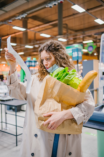 Young woman is unpleasantly surprised while looking into a paper check in a grocery supermarket and holding a shopping bag. Rising food prices.