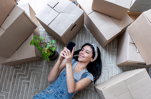 Beautiful young woman taking a break from unpacking boxes lying down on the floor while looking at videos on smartphone smiling - New beginnings concepts