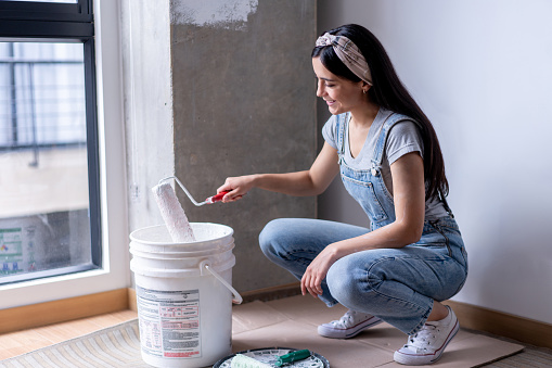Latin American beautiful young woman at her new flat painting a wall with white paint and using a roller - DIY concepts
