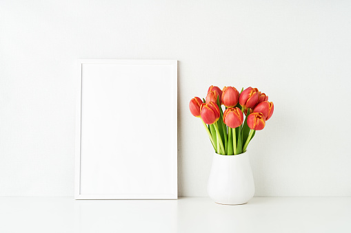 White frame mockup and red tulips in a white vase on a white table by the wall. Copy space for text