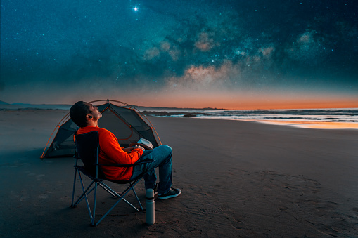 man sitting in a camping chair alone on the beach looking at the starry night next to a tent