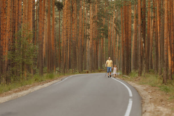 A young man father in shorts and a T-shirt walks through a beautiful pine forest holding hands with his daughter in a dress and hat. The concept of family walks, fatherhood.