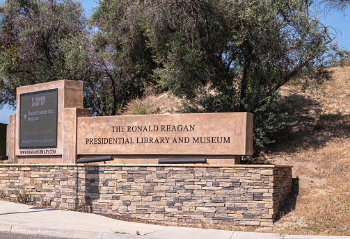 Simi Valley, California, USA - April 27, 2022: Ronald Reagan Presidential Library. Brown stone sign and electronic display at road entrance of domain. Green foliage in back. Patches of blue sky.