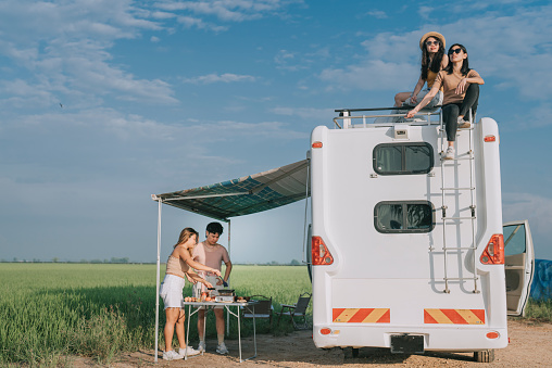 Asian Chinese Lesbian couple enjoying scenic view on top of campervan while friends cooking breakfast outdoor road trip during weekend morning