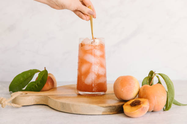 Mixing a fresh peach drink in a cup of ice A human hand is mixing a peach drink with a cold spoon. In the drink is also water and ice. There are peaches, some whole and some cut up. The drink is on a wood cutting board in a marble kitchen. iced tea stock pictures, royalty-free photos & images