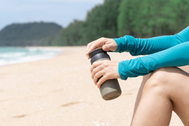 Closeup of womans hands holding reusable water bottle sitting at the beach on sunny day. Closeup of womans hands holding insulated stainless reusable water bottle sitting in on beach on sunny day, living eco friendly, zero waste and green living lifestyle. asian drinking water while on travel stock pictures, royalty-free photos & images