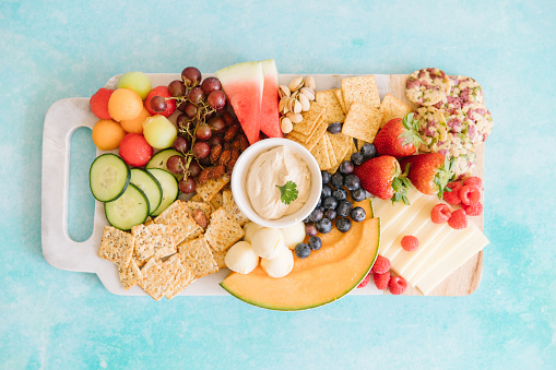 A bright charcuterie board on a blue background. On the charcuterie board are grapes, watermelon, cantaloupe, crackers, grapes, cheese, strawberries, hummus, cucumber, pistachios, blueberries on a marble slab.