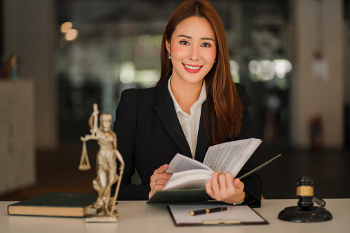 Attractive Asian Lawyer Female Lawyer Discussing Contract Documents with Golden Goddess Scales with Hammer on Wooden Tables in Office of Legal Services, Advice, Justice and Real Estate Ideas.