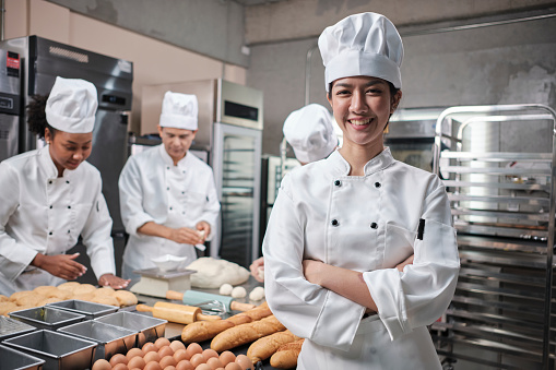 Young beautiful Asian female chef in white cooking uniform looks at camera, arms crossed and cheerful smile with food professional occupation, commercial pastry culinary jobs in a restaurant kitchen.