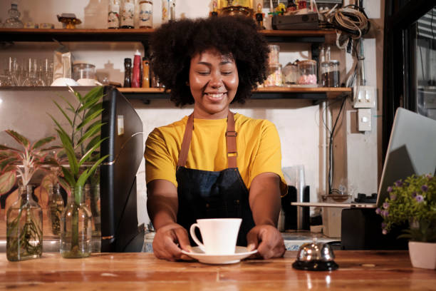 African American female barista cheerful offers a cup of coffee in casual cafe. stock photo