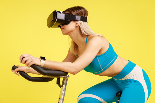 Side view portrait of fit grinning woman in blue sportswear exercising on sports machine wearing VR-headset, having cardio workout. Indoor studio shot isolated on yellow background.